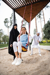 Canvas Print - Happy arabian girl swinging on playground near parents in park