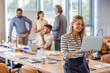 © Dragana Gordic - Woman manager holding laptop and standing in modern office. Businesswoman in casual wear holding laptop smiling and looking to camera while her colleagues communicating in the background
