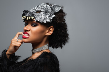 stylish african american woman in werewolf halloween mask touching lips while grinning isolated on grey