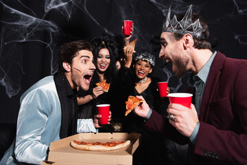 Wall Mural - amazed men looking at friend near pizza and excited interracial women in halloween costumes on black