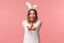 Holidays, Spring And Party Concept. Portrait Of Lovely, Romantic Young Blond Woman In Rabbit Ears And White Dress, Giving You Painted Easter Eggs As Celebrating Orthodox Holiday, Pink Background
