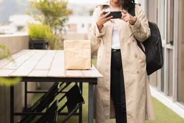 Wall Mural - Young woman in a cream coat with a phone and a paper lunch bag.