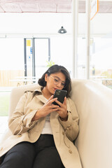 Wall Mural - Vertical shot of the young woman in a cream coat lying on the couch and looking at the phone.