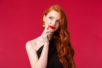 Wall Mural - Close-up portrait of dreamy elegant and stylish redhead woman in black dress, red lipstick, making plan, thinking and smiling pleased of finding great idea, touch lip and look away pondering