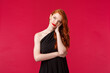 Waist-up portrait of annoyed and bored, bothered redhead woman in elegant black dress, sighing displeased, getting pressured, look up troubled and upset, stand red background