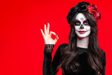Girl With Creative Sugar Skull Makeup With A Wreath Of Flowers On Head, Wide Isolated On Red Background. Holiday Concept Dia De Los Muertos  On Poster For Halloween Party Or La Calavera Catrina.