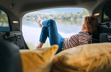 Woman Dressed In Warm Calm Colors Clothes And Jeans Lying In The Cozy Car Trunk And Enjoying The Mountain Lake View. Warm Early Autumn Auto Traveling Concept Image.