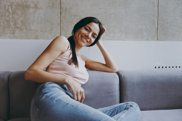 Wall Mural - Front view smiling magnificent happy young latin woman 20s in casual clothes sit on sofa look camera rest relax spend time in living room in own house be lost in reverie good day. Real estate concept.