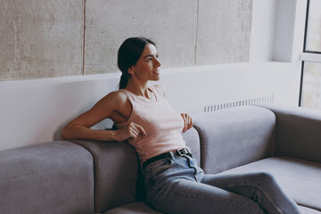 Wall Mural - Side view profile excited vivid young latin woman 20s in casual clothes sit on sofa enjoy free time rest relax spend time in living room in own house be lost in reverie good day. Real estate concept.