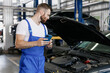 © ViDi Studio - Young professional technician car mechanic man 20s in blue overalls white t-shirt use hold mobile cell phone surfing fix problem with raised hood bonnet work in vehicle repair shop workshop indoor