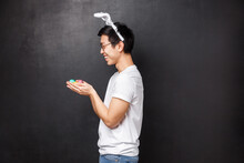 Holidays, Party And Easter Concept. Profile Portrait Of Cheerful Cute Young Asian Man In Rabbit Ears And White T-shirt, Standing Over Black Background With Painted Eggs In Hands