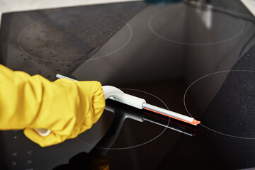 Cleaning induction stove. Woman in yellow rubber gloves cleans kitchen induction hob with cleaning sparay. Clear kitchen appliance