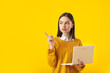 © Pixel-Shot - Beautiful young woman with laptop pointing at something on yellow background