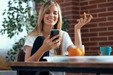 Wall Mural - Smiling young woman enjoying breakfast while using her mobile phone in the living room at home.