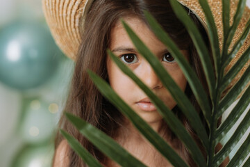 Wall Mural - Face of a little girl surrounded by tropical leaves. Closeup portrait of a beautiful swarthy baby with perfect skin and dark hair. Natural cosmetics, health, cleanliness, skin care, beauty concept