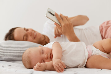 Happy smiling with with pleasant appearance wearing white casual style t shirt using cell phone, looking at device screen with positive emotions, lying on bed with her little sleeping daughter.
