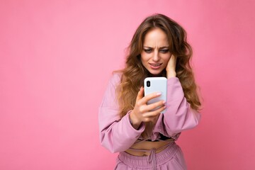 Beautiful amazed surprised blonde young woman wearing pink hoodie isolated over pink background with copy space holding and using mobile phone reading sms looking at device display and don't