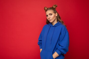 Wall Mural - Portrait of dissatisfied offended young attractive winsome blonde woman with two horns with sincere emotions wearing casual bright blue hoodie isolated on red background with free space