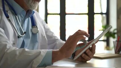 Senior Doctor Thinking About Patient's Diagnosis and Typing on a Tablet Computer. His Office is Bright and Modern.