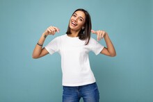 Portrait Of Positive Happy Smiling Young Beautiful Brunette Woman With Sincere Emotions Wearing Casual White T-shirt For Mockup Isolated Over Blue Background With Copy Space And Pointing At Empty
