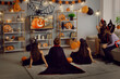 © Studio Romantic - Kids in spooky costumes of witches, monsters and pirates watching children's Halloween movie sitting on floor and sofa in interior decorated with orange and black pumpkins at fancy dress party at home