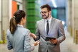 © dusanpetkovic1 - Warehouse manager training. A man and a woman in a business suit stand in a warehouse and talk about work. A smiling man with glasses holds a tablet in his hand and looks the woman straight in the eye