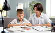 © MYDAYcontent - Caucasian schoolboy sitting with mother at desk and reading book. Caring woman helping son with homework. Educational moments during parenting.