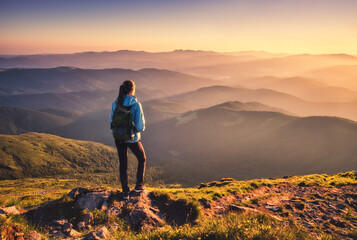 Girl with backpack on mountain peak with green grass looking in beautiful mountain valley in fog at sunset in autumn. Landscape with sporty young woman, foggy hills, orange sky in fall. Hiking. Nature