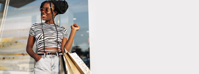 Happy black young woman walking in the mall after shopping for clothes, looking out the store window