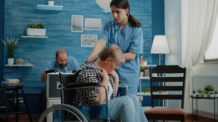 Nurse using stethoscope for heartbeat checkup on disabled woman in nursing home. Senior patient with disability in wheelchair receiving consultation from medical assistant for recovery