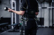 © Dusan Petkovic - A portrait of a body part of a woman in a black EMS training suit standing in the middle of a gym. She holds dumbbells in her hands and does arm training. Contemporary coaching concept