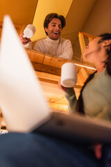 Wall Mural - Low angle view of smiling man with cup looking at blurred girlfriend with laptop in glamping house