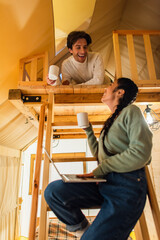Wall Mural - Low angle view of smiling interracial couple with cups and laptop looking at each other in glamping house