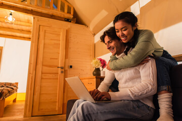 Wall Mural - Smiling asian woman hugging boyfriend with laptop on couch in glamping house