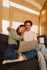 Wall Mural - Smiling multiethnic couple with cup and laptop sitting on couch in glamping house
