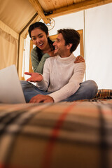 Wall Mural - Smiling man pointing at laptop near asian girlfriend in glamping house