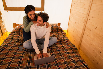 Wall Mural - Smiling asian woman hugging boyfriend using laptop on bed in glamping house