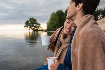 Wall Mural - smiling asian woman with closed eyes holding cup near boyfriend and lake