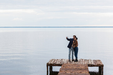 Wall Mural - happy multiethnic couple taking selfie on cellphone on pier near sea