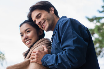 Wall Mural - Low angle view of man with closed eyes hugging asian girlfriend in blanket outdoors