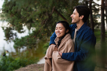 Wall Mural - Smiling man hugging asian girlfriend in blanket outdoors