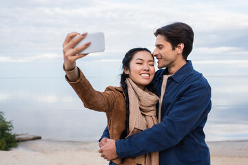 Wall Mural - smiling man hugging asian girlfriend taking selfie on beach