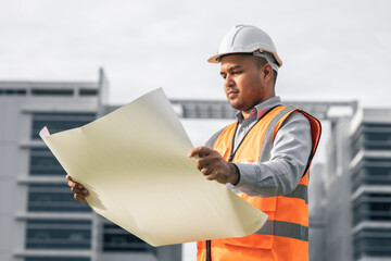 Asian engineer handsome man or architect open the paperwork blueprint and planning to building construction. Worker asian man with white safety helmet working in modern building construction site.