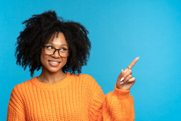 Cheerful african woman in glasses looking up and pointing finger to empty space for product, online courses or service advertising. Young black woman casual student gesturing showing place for ads