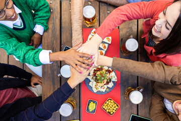 Millennial group of united friends stacking hands - Happy young multiracial students gathering while having fun and celebrating together at brewery bar - Teamwork, community and unity concept