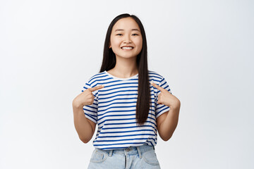 Portrait of pretty asian woman pointing fingers at herself and smiling pleased, self-promoting, introduce her, standing over white background