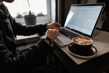 professional man with a luxury watch in a black hoodie works on a laptop and drinks coffee at a table in a cafe