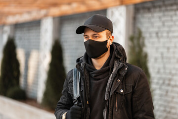 Wall Mural - Young man with a black protective mask and a fashionable cap in a black stylish hoodie and a winter jacket with a backpack walks in the city