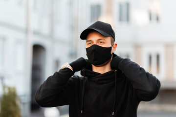 Wall Mural - Fashionable young man with a protective medical mask in a black stylish hoodie with a fashion cap on the street. Modern male urban style and pandemic concept