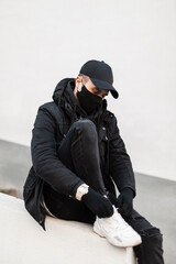 Wall Mural - stylish man model with medical mask and cap in fashion urban clothes look with jacket. hoodie and sneakers sits and ties his shoelaces on the street